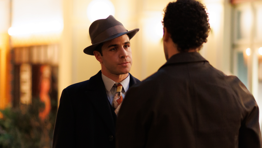 Tenso encuentro entre dos hombres: uno en traje oscuro y sombrero mira fijamente a otro de espaldas, en un espacio público con iluminación cálida.