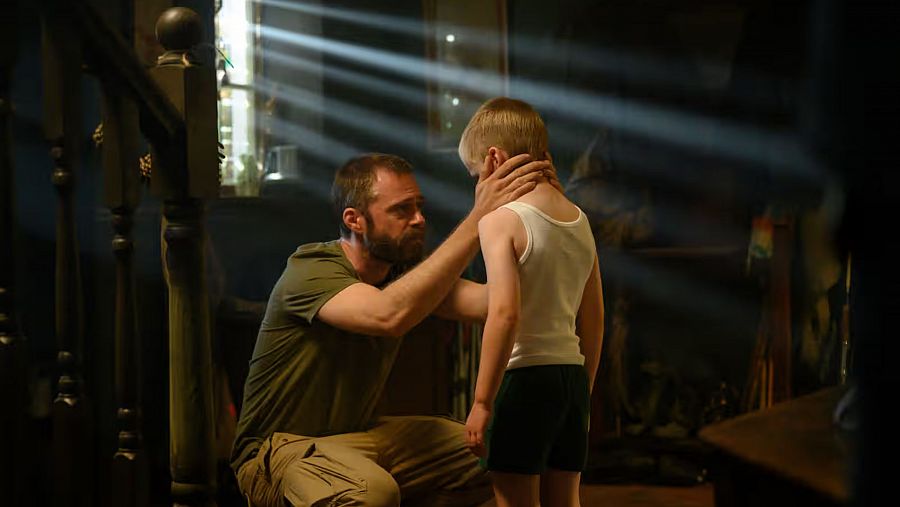 Hombre y niño en habitación oscura, cerca de escalera de madera. Iluminación dramática, atmósfera tensa. Posible escena de serie RTVE Play.