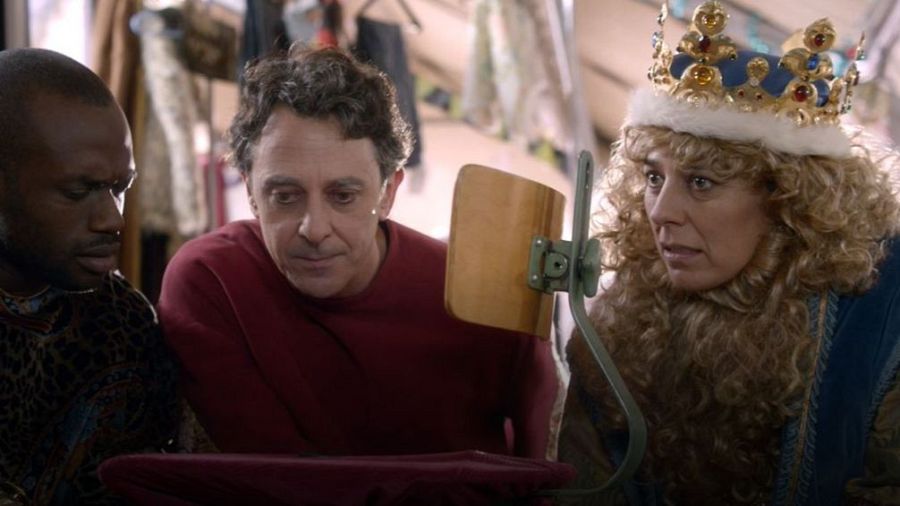 Detrás de cámaras de una película navideña. Una mujer con corona azul y dos hombres observan algo juntos. Vestuario y equipo de rodaje visibles.