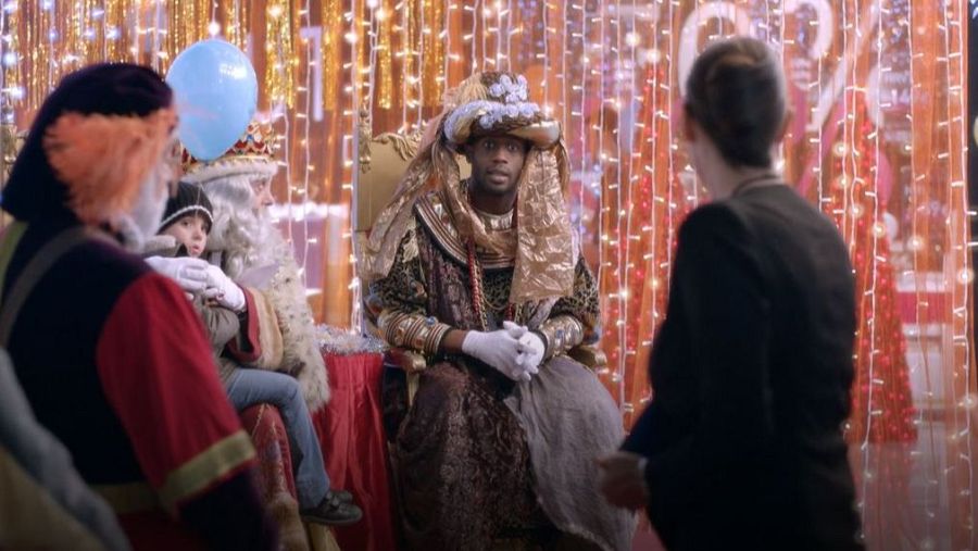 Escena navideña con Djédjé Apali como Rey Mago en un trono dorado, junto a Papá Noel, un niño y una mujer. Iluminación festiva.