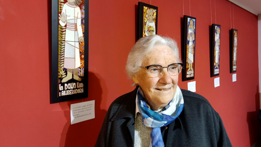 Regina Goberna sonríe junto a sus obras de arte: paneles con figuras estilizadas, posiblemente cerámicas, con una etiqueta que dice 