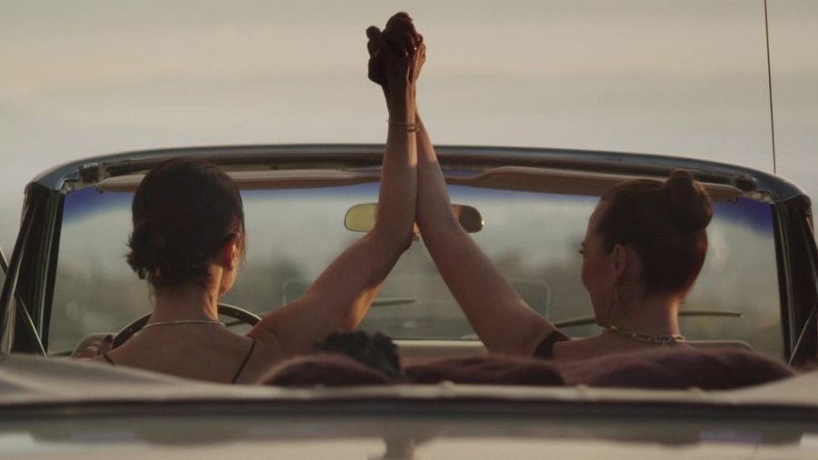 Dos mujeres en un descapotable clásico, con el pelo recogido y manos entrelazadas sobre el parabrisas, celebran con alegría al atardecer.