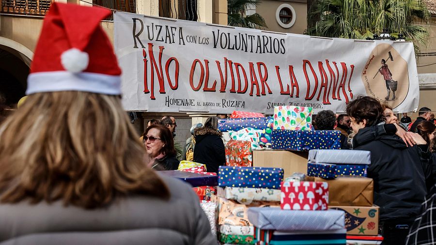 Regalos de varios tamaños y colores son distribuidos por voluntarios. Una pancarta indica un homenaje en Ruzafa el 9 de noviembre, posiblemente por una DANA. Se observa un gorro de Papá Noel.