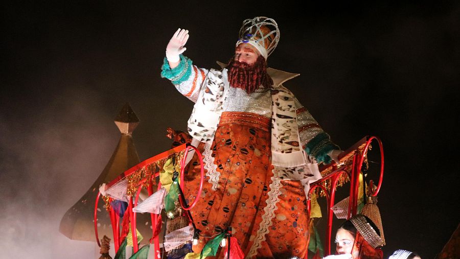 Rey Gaspar en cabalgata, Barcelona.  Túnica blanca y naranja, corona, barba roja. Saluda desde carroza decorada con humo artificial.