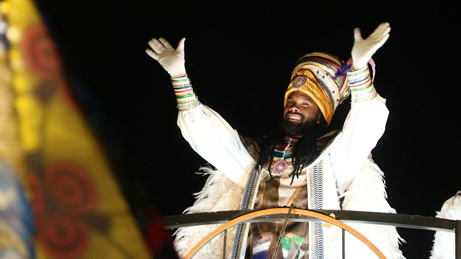 Rey Gaspar saludando desde su carroza en una cabalgata nocturna. Luce túnica blanca dorada, tocado alto y guantes. Fondo oscuro.