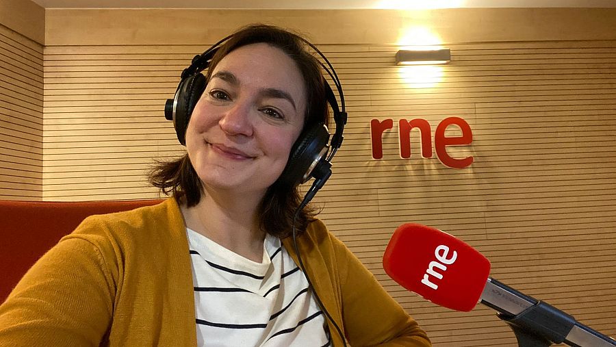 Mujer sonriente en estudio de radio RNE, con auriculares y chaqueta mostaza, frente a un micrófono con el logo RNE. Iluminación cálida, pared de madera.