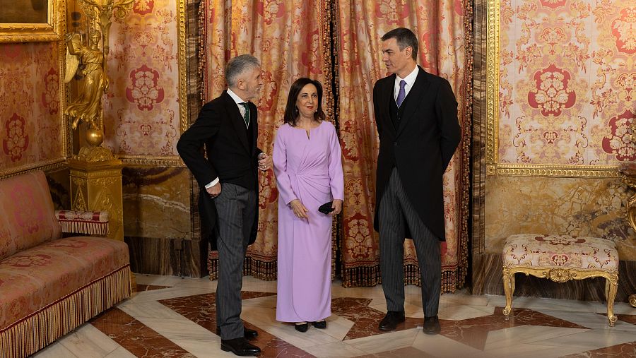 Tres personas en un salón clásico, con decoración lujosa. Uno viste traje oscuro, otro un vestido largo lila, y el tercero también traje oscuro.