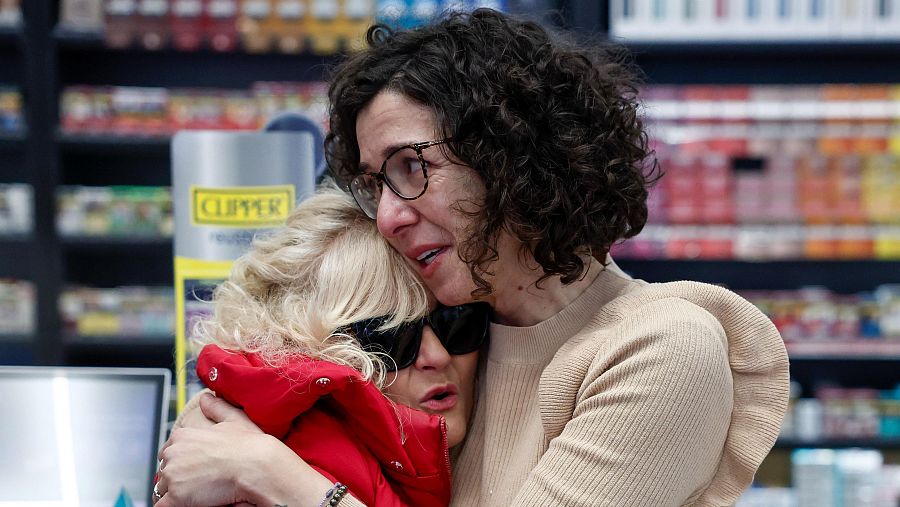 Abrazos de alegría entre dos mujeres en un local con estanterías y un cartel amarillo con la palabra 