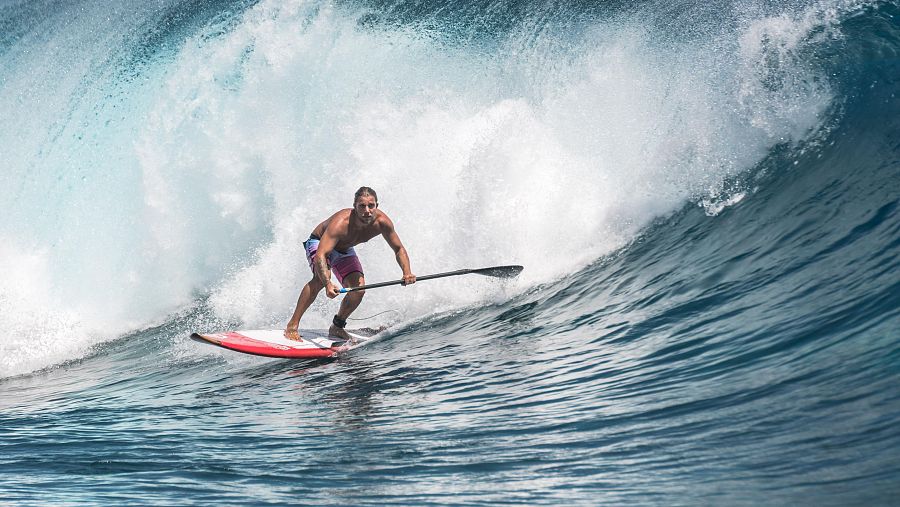 Surfista con coleta y pantalones cortos sobre tabla roja y blanca, navegando una gran ola de océano azul oscuro.