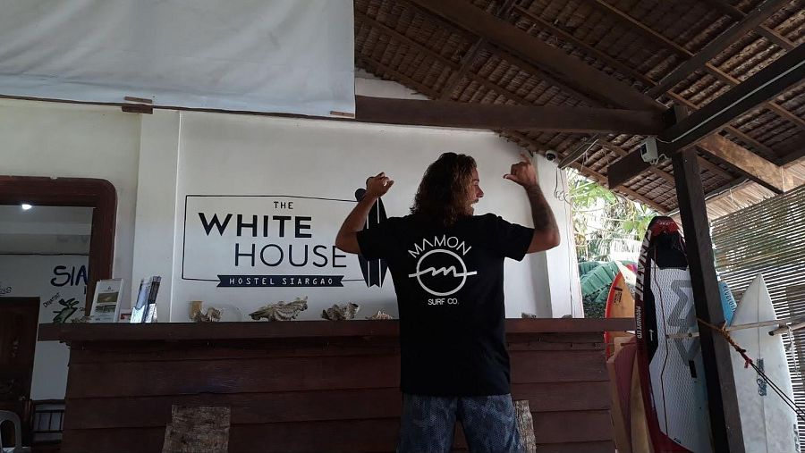 Diego Bello muestra su aprobación con los pulgares arriba en un albergue con tablas de surf. Lleva una camiseta negra con el logo 