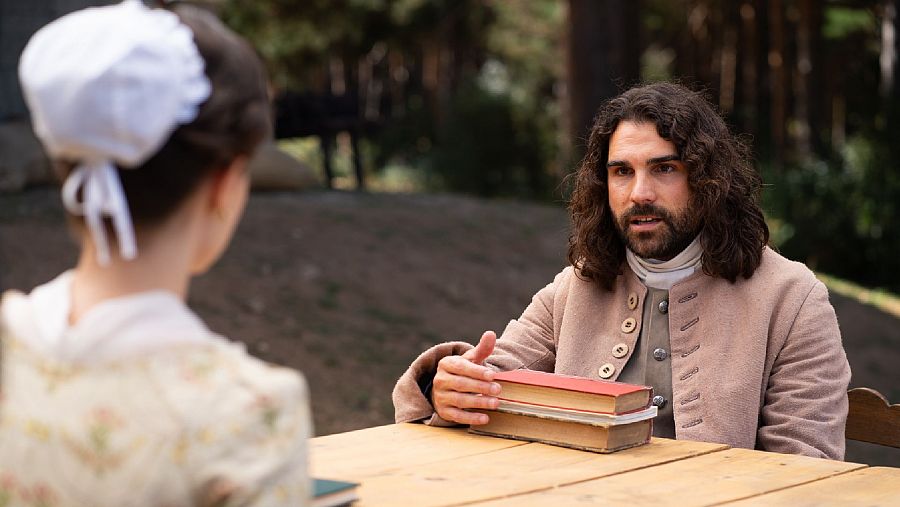 Hombre y mujer, vestidos de época, conversan a una mesa con libros antiguos en un entorno natural.  Posible escena de 