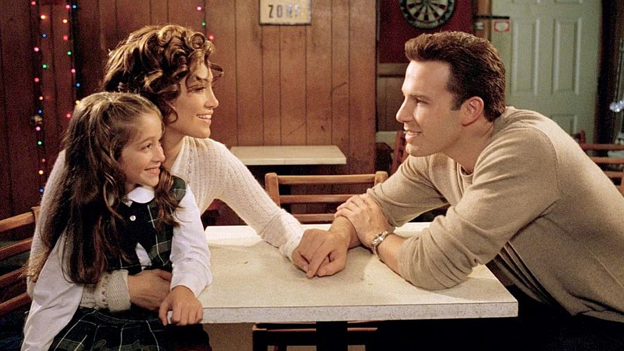 Jennifer Lopez, Ben Affleck y una niña, sentados juntos en una mesa, en una escena cálida y familiar de una película.  Ambiente decorado con luces navideñas.
