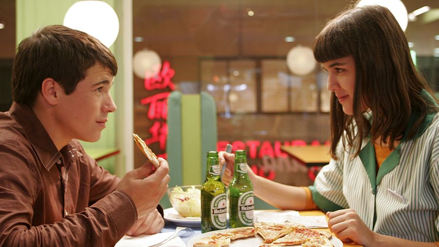 En un restaurante, un hombre con camisa marrón y una mujer con uniforme a rayas comparten pizza y cerveza Heineken.  Escena informal de la película 