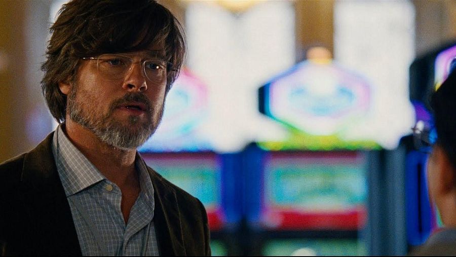 Brad Pitt, con gafas y camisa a cuadros, en una escena ambientada en un casino.  Expresión seria, pelo castaño y barba.  Parte de otra persona visible.