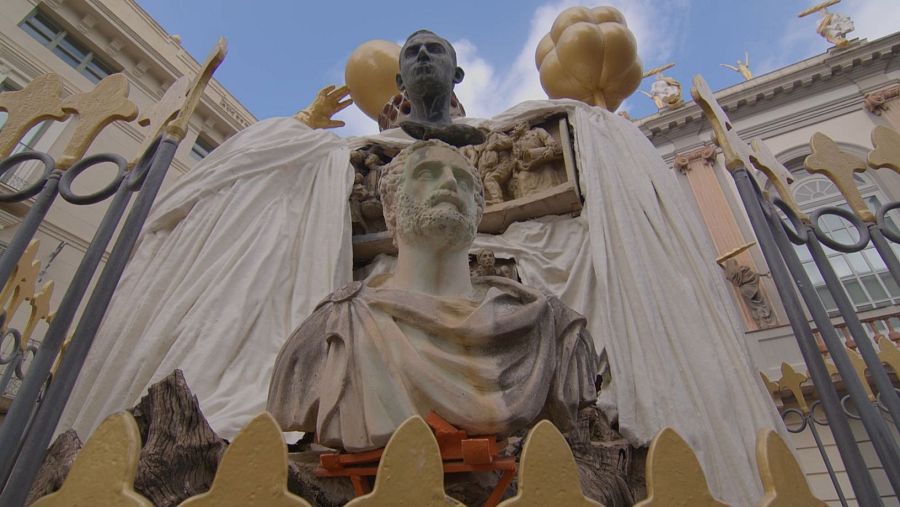 Escultura con dos bustos, uno principal con barba y otro pequeño encima, flanqueada por objetos dorados y cubierta parcialmente por una tela blanca. Vista desde detrás de una valla.