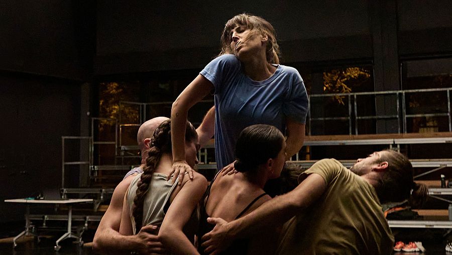 Intensa escena de una obra de teatro: cinco personas, una mujer en el centro sostenida por otras cuatro, en un momento de gran esfuerzo físico y emocional. Iluminación dramática.