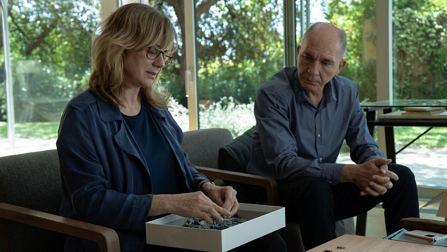 Mujer con gafas y chaqueta azul resuelve un puzzle; hombre calvo con camisa azul la observa.  Interior luminoso, tensión contenida.