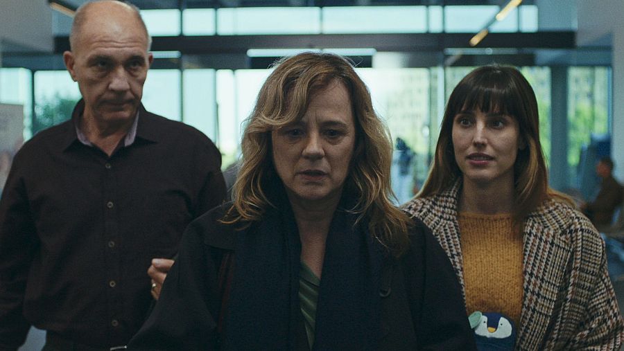 Tres personas con expresiones serias en un interior luminoso.  La mujer de mediana edad parece angustiada.  El vestuario incluye camisa oscura, abrigo oscuro y abrigo de cuadros.