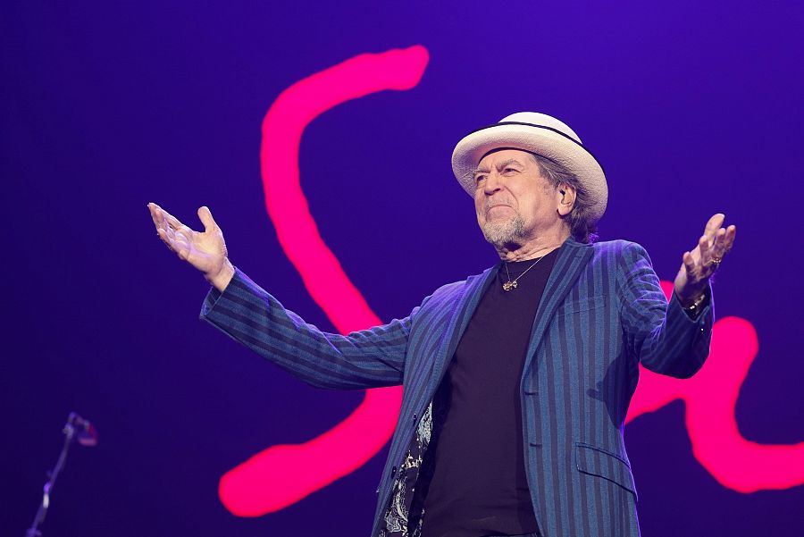 Joaquín Sabina en concierto: sombrero blanco, chaqueta azul marino a rayas, camiseta negra, gesto de saludo, fondo morado oscuro con una gran 'S' rosa.