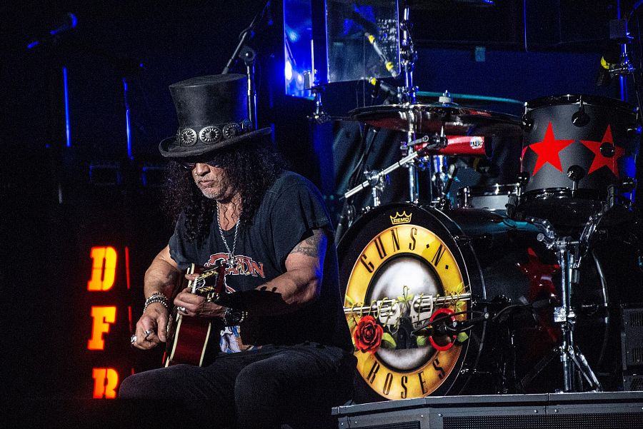 Slash, con sombrero y gafas, toca una guitarra acústica en un escenario con iluminación oscura y el logo de Guns N' Roses visible en la batería.