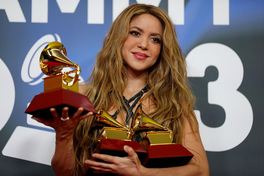 Shakira muestra tres gramófonos dorados de los Latin Grammy. Lleva un top oscuro y un anillo. Su cabello rubio cae en ondas sobre sus hombros.