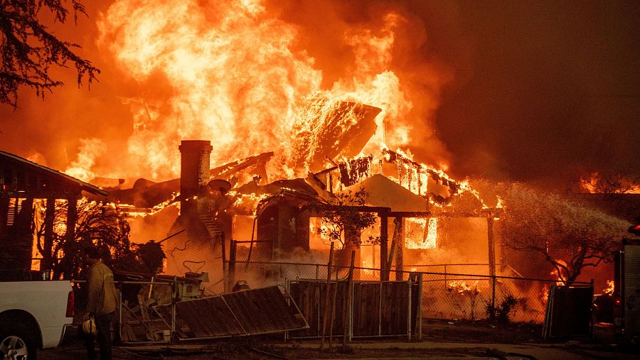 Intenso incendio consume una casa de madera; bomberos con equipo de extinción trabajan en la escena.