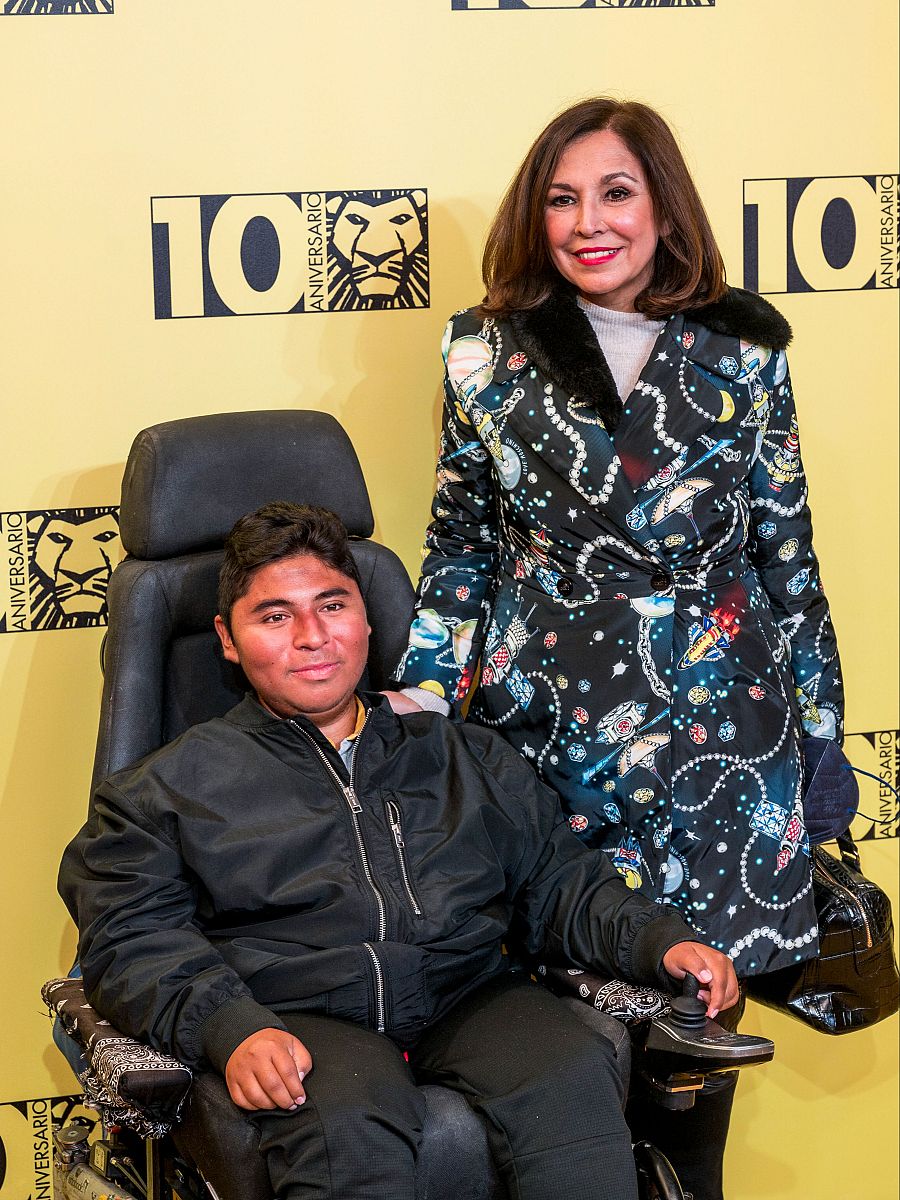 Fotografía de Isabel Gemio y Gustavo en un evento de aniversario. Él en silla de ruedas con chaqueta negra; ella con abrigo negro estampado y cuello de pelo.