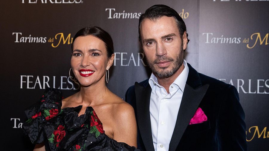 Arantxa del Sol, con vestido negro y pendientes grandes, y Finito de Córdoba, con traje oscuro y pañuelo fucsia, posan en un evento con fondo promocional que incluye las inscripciones 