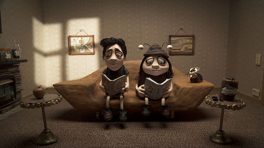 Dos personajes de plastilina leen sentados en un sofá, en una sala de estar con chimenea, mesas y cuadros.  Ambiente cálido y tranquilo.