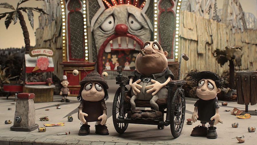 Escena de animación con tres figuras de plastilina: un adulto en silla de ruedas y dos niños con semblante preocupado, en un parque de atracciones descuidado y sucio.