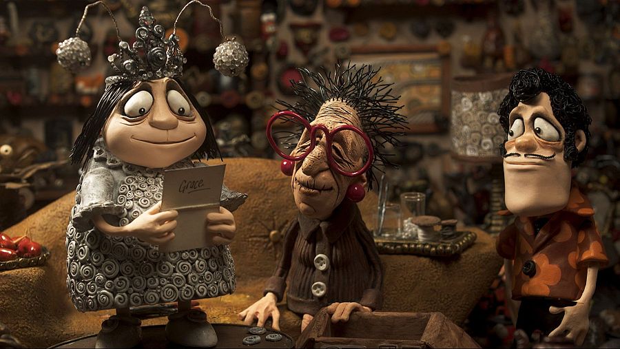 Escena de animación stop-motion con tres personajes: mujer con vestido gris y carta, mujer con gafas rojas y hombre con camisa naranja. Ambiente hogareño.