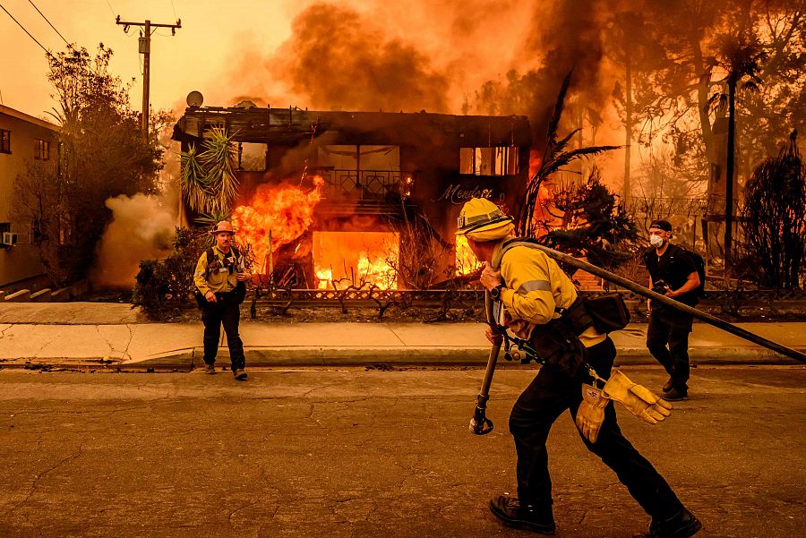 Bomberos combaten un incendio en un edificio de apartamentos envuelto en llamas y denso humo naranja.  Se observa a un bombero con manguera corriendo hacia el edificio.