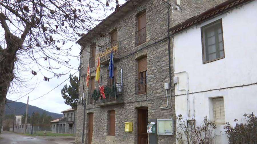 Ayuntamiento de piedra de dos plantas con balcones, banderas de España y UE, corona navideña y buzón amarillo en la fachada.  Calle y montañas al fondo.