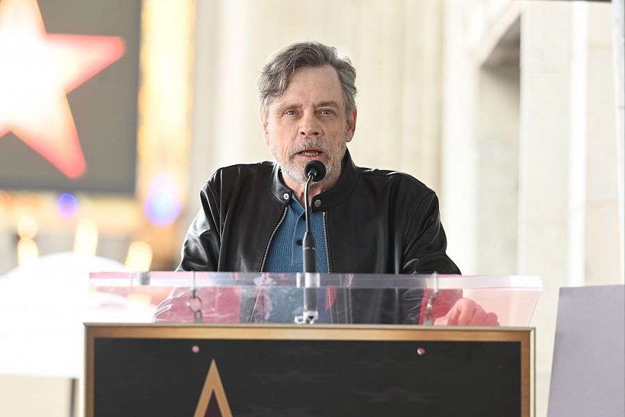 Actor con chaqueta negra y camisa azul marino da un discurso tras un atril de madera con superficie acrílica transparente; al fondo, una estrella roja borrosa.