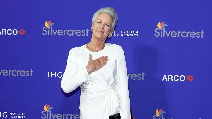 Actriz con vestido blanco y cabello corto gris, posando ante un fondo azul con logotipos de patrocinadores.