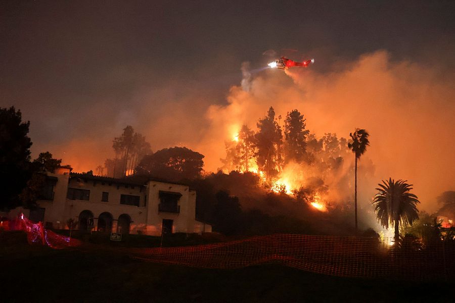 Incendio forestal nocturno con llamas intensas, un helicóptero en acción y una casa cercana protegida por una malla naranja. El fuego ilumina palmeras en la escena.