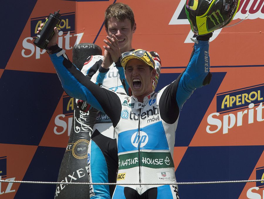 Piloto de motociclismo celebrando una victoria en el podio, con mono blanco y azul, botella de champán y casco en mano. Fondo naranja con patrocinadores.