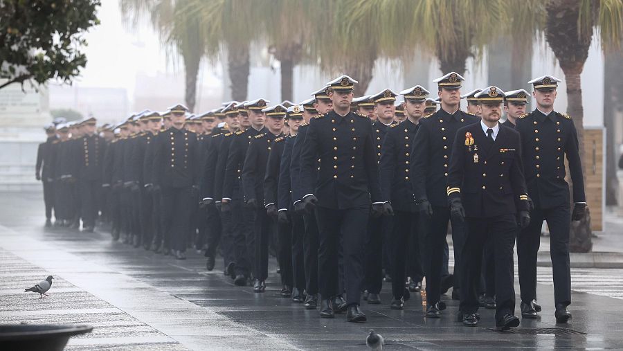 Guardamarinas en uniforme de gala marchan por una calle mojada, con árboles y palmeras al fondo. Se observan dos palomas.