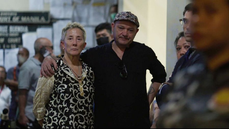 Un hombre y una mujer, con expresiones serias, abrazados en un juzgado. Ella viste una blusa con estampado de arabescos; él, camisa negra y gorra.  Fondo con gente, algunas enmascarilladas.