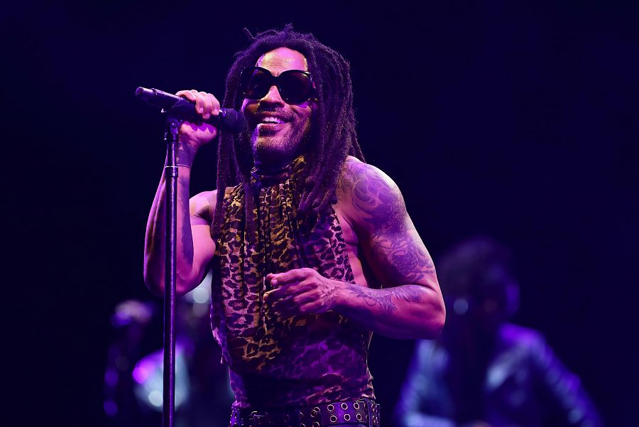 Cantante con gafas de sol y camiseta estampada actuando en un escenario con iluminación púrpura, sosteniendo un micrófono y con la mano levantada hacia el público.