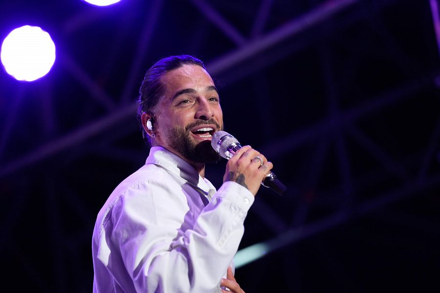 Cantante con camisa blanca y micrófono, actuando en un escenario con iluminación púrpura.  Pelo recogido y auricular visibles.