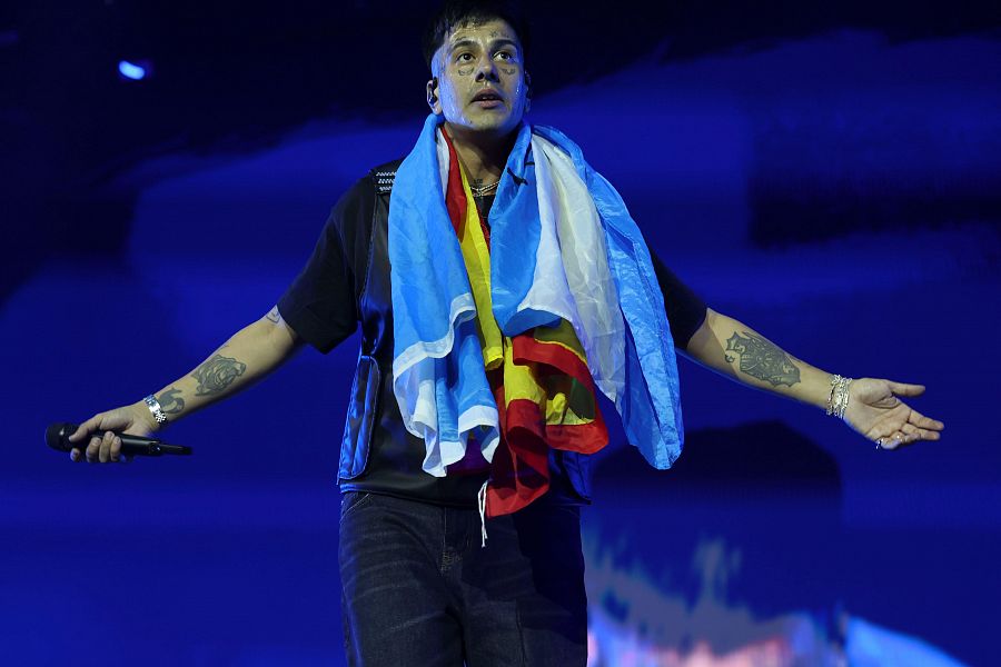 Cantante en concierto con micrófono, camiseta negra y pantalones oscuros.  Banderas de Argentina y España al cuello. Varios tatuajes visibles.