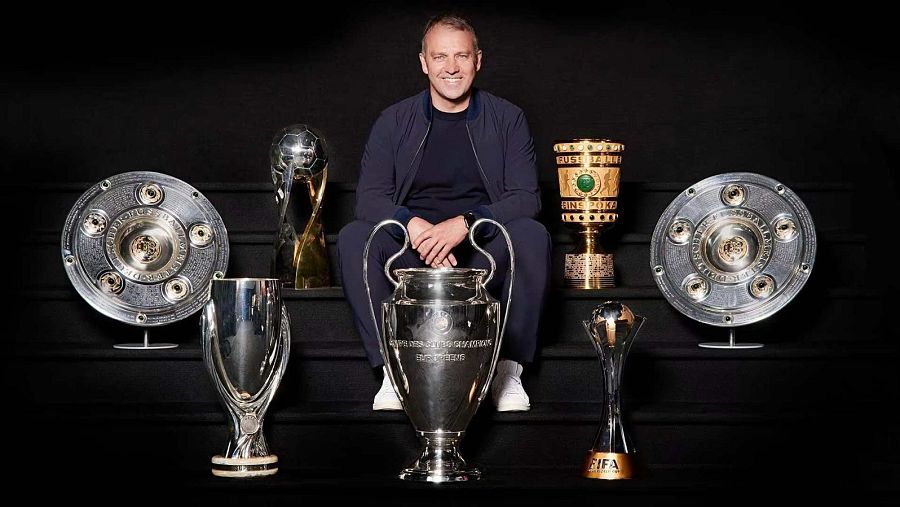 Hansi Flick, sentado en escaleras, posa con los seis trofeos del sextete del Bayern de Múnich (2019-2020), incluyendo la Champions League.