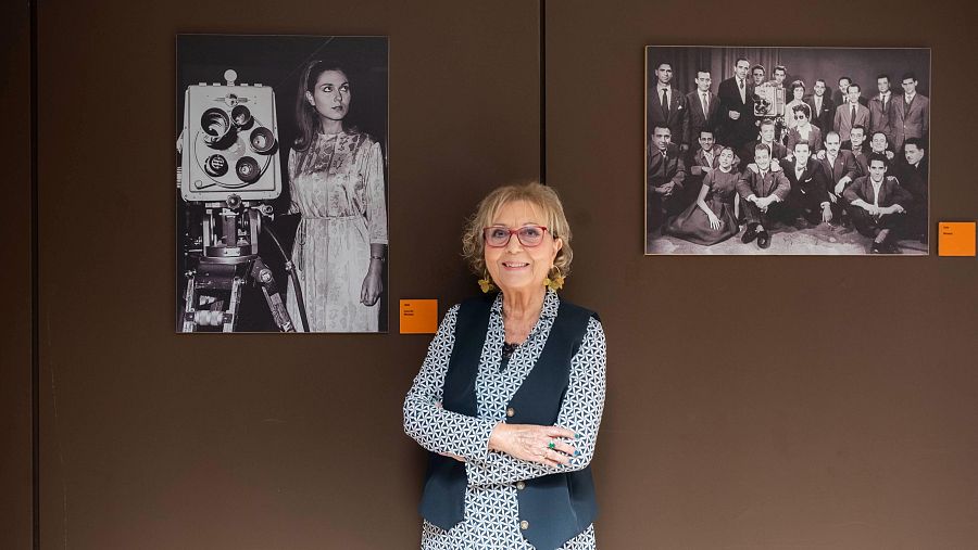 Mujer mayor con gafas y chaleco oscuro sonríe ante dos fotos antiguas en blanco y negro; una muestra a una mujer joven con una cámara de televisión, la otra a un grupo en ropa formal.