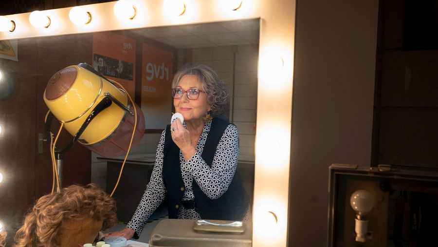 Mujer con gafas y chaleco oscuro maquillándose frente a un espejo con bombillas, junto a un secador de casco amarillo y una peluca.