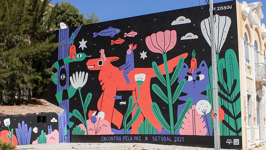 Mural con perro naranja, personajes azules, gato morado y diversas plantas y animales sobre fondo negro. Firma 