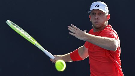 Tenista en acción golpeando una pelota amarilla; viste camiseta roja y gorra blanca con logo.  Fondo: pista azul.