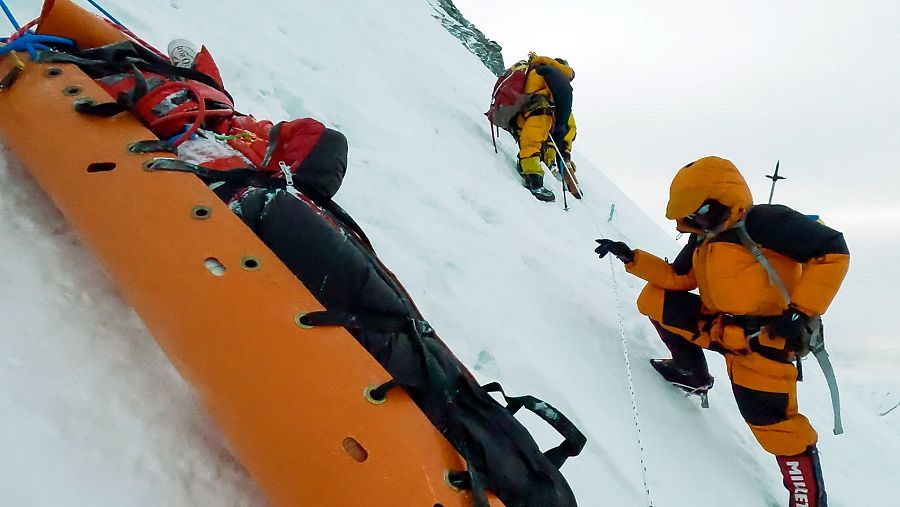 Rescate en alta montaña: alpinista en camilla naranja siendo descendido por una ladera nevada, asistido por dos rescatistas.