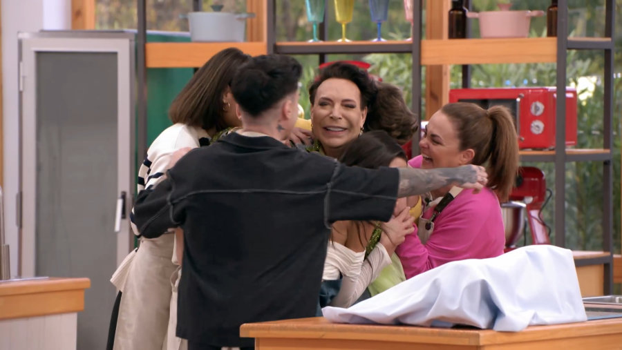 Personas abrazándose en una cocina, una con cabello oscuro sonríe en el centro.  Utensilios de cocina y un horno microondas rojo se ven al fondo.