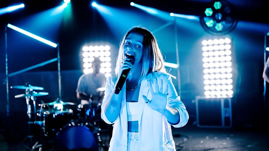 Cantante con chaqueta blanca actuando en un escenario iluminado, con batería visible al fondo, durante una presentación en directo.